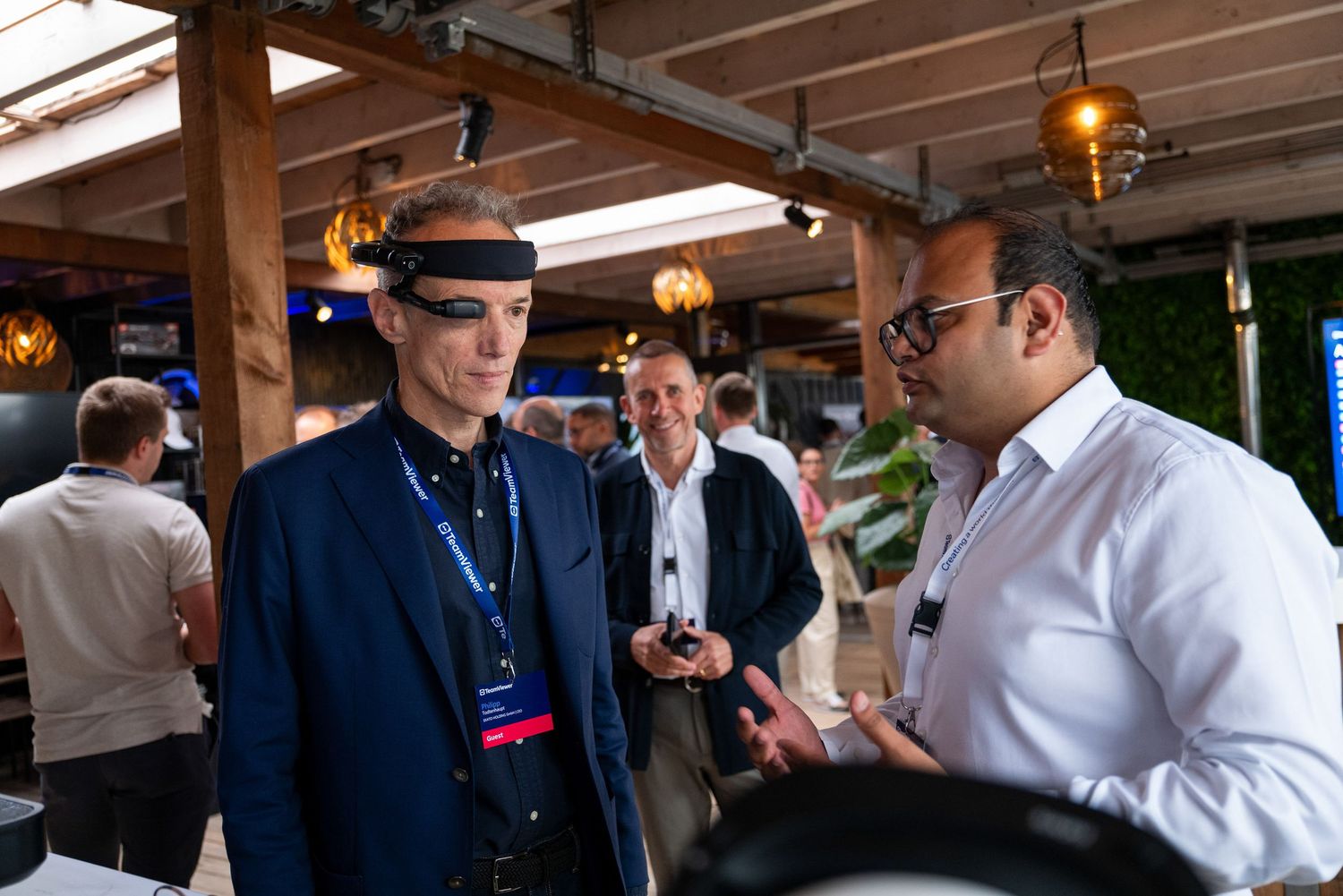 Business professionals engaged in conversation at a corporate event with wooden ceiling beams visible.