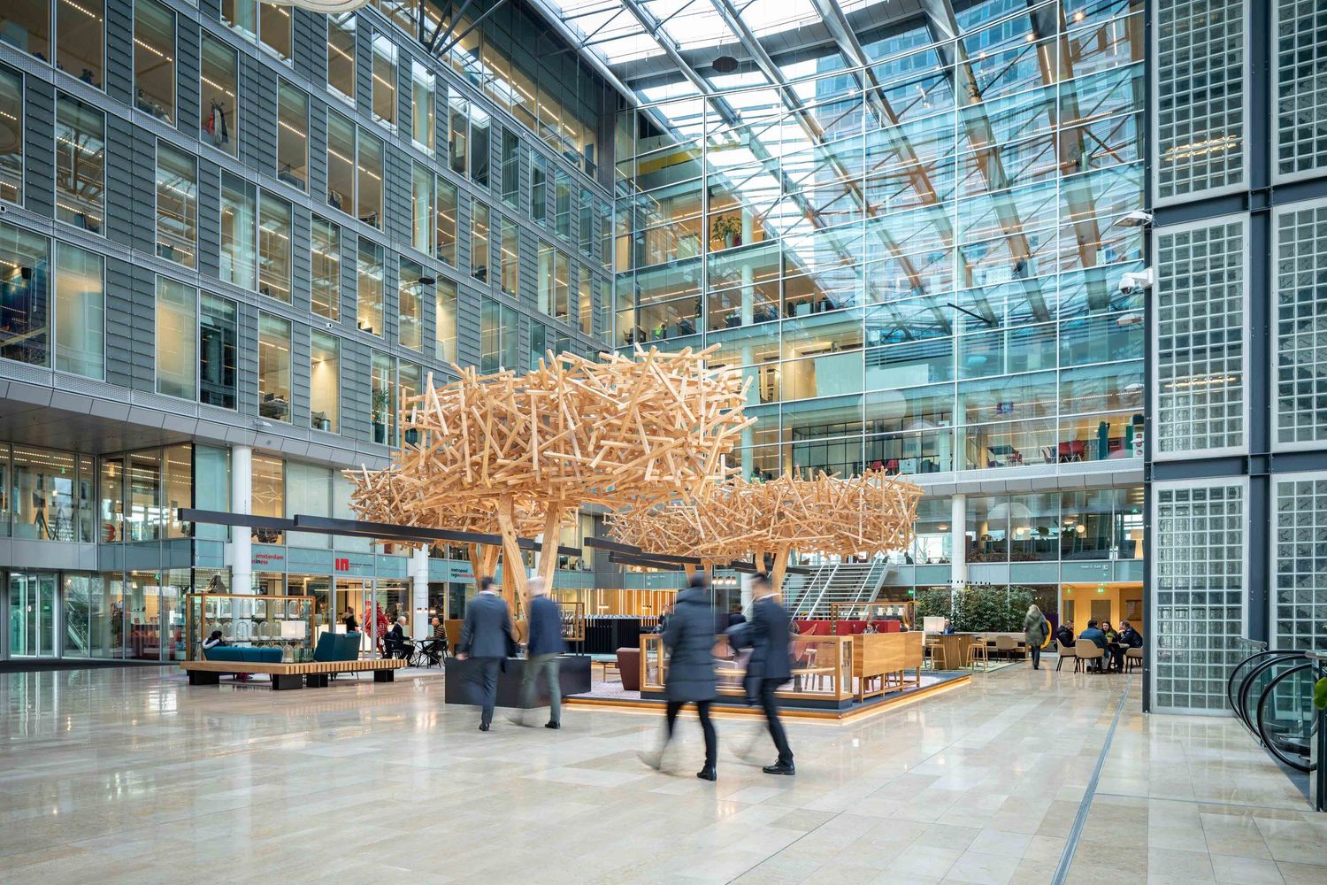 Modern office atrium with glass ceiling and large wooden art installation in the center of a busy lobby space.