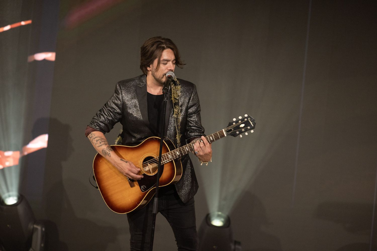 A musician performs on stage with an acoustic guitar under dramatic lighting during a live concert performance.