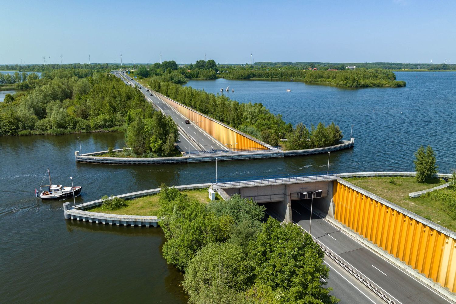 Aerial view of a modern aqueduct bridge over a lake in the Netherlands, with yellow barriers and a boat passing below.