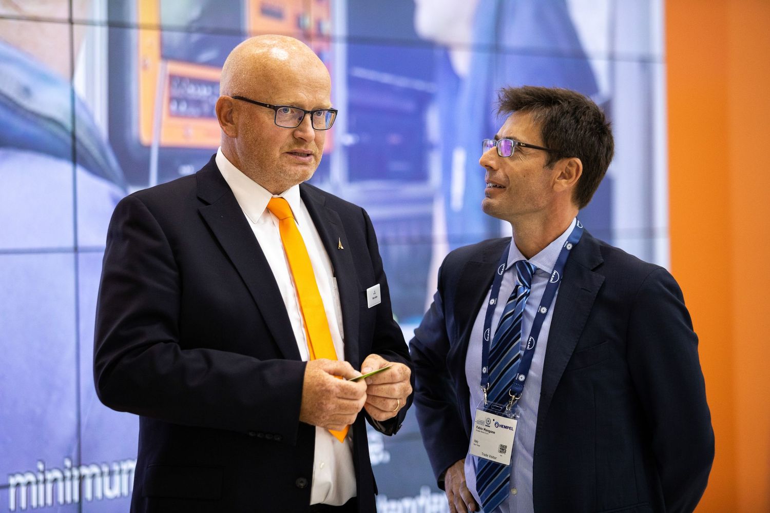 Two people in business attire having an animated conversation at a professional event with orange walls.