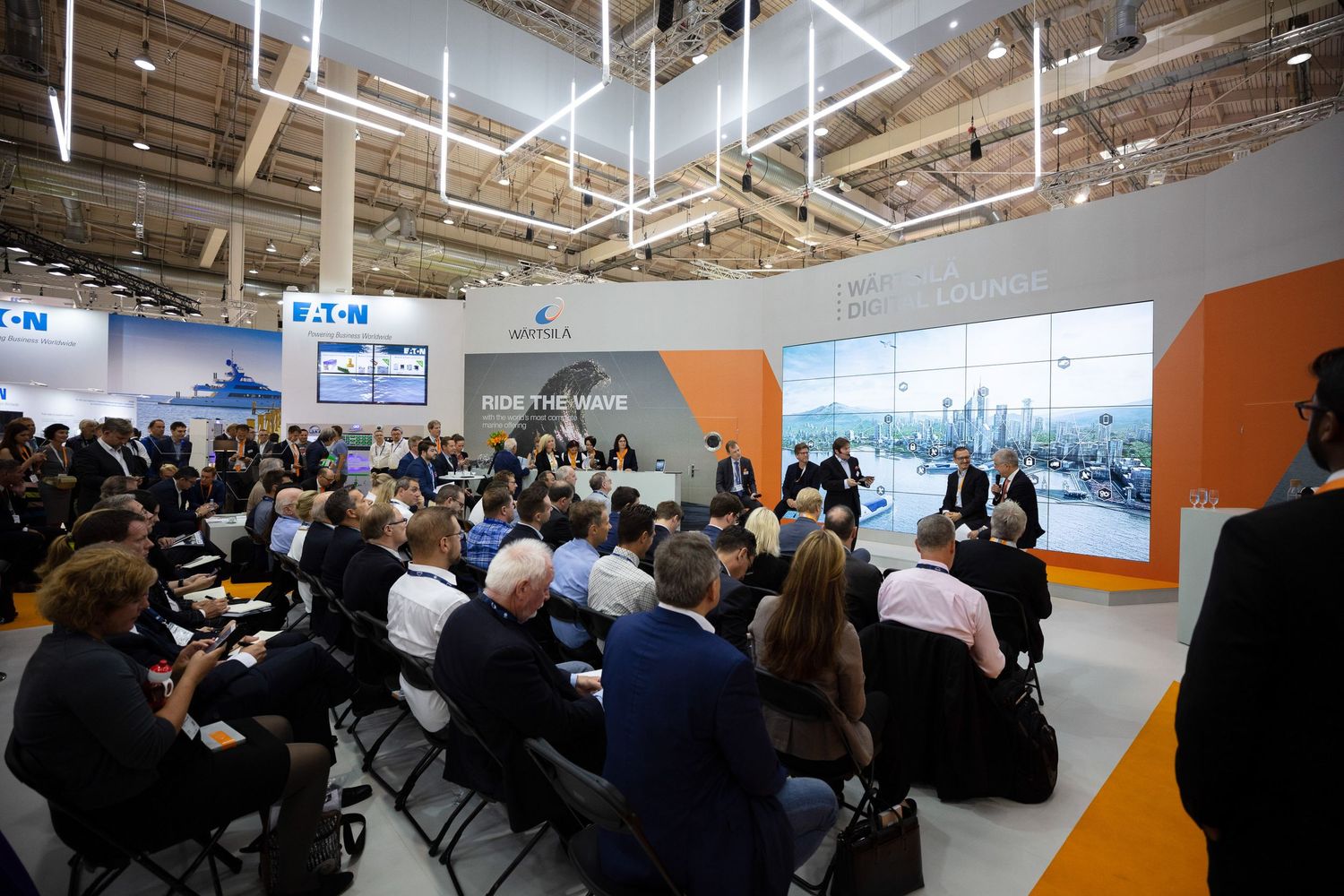 Large audience seated in rows at a modern office space with exposed ceiling and glass walls during a presentation.