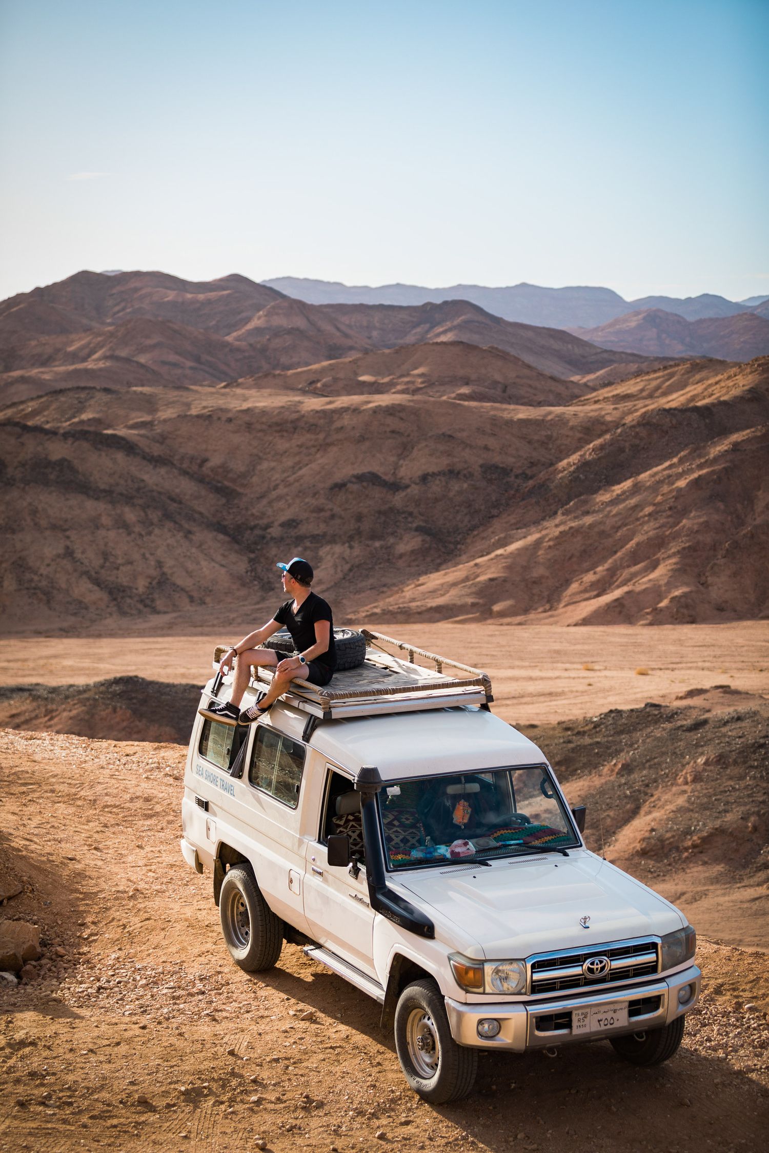 White pickup truck driving through rugged desert mountain terrain with adventurous passengers on the roof.