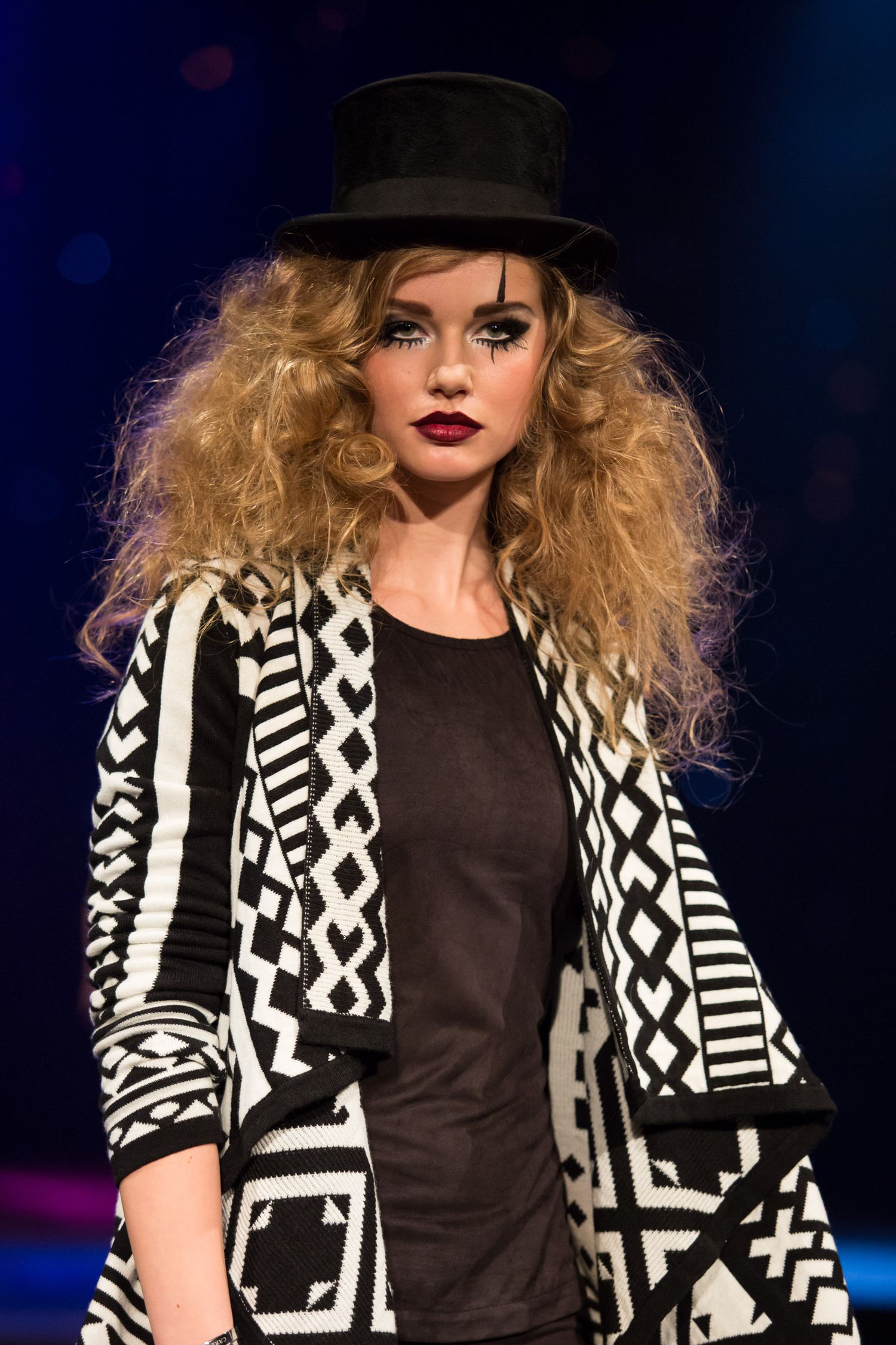Model wearing black top hat, curly blonde hair, dramatic makeup, and black and white geometric patterned cardigan on runway.