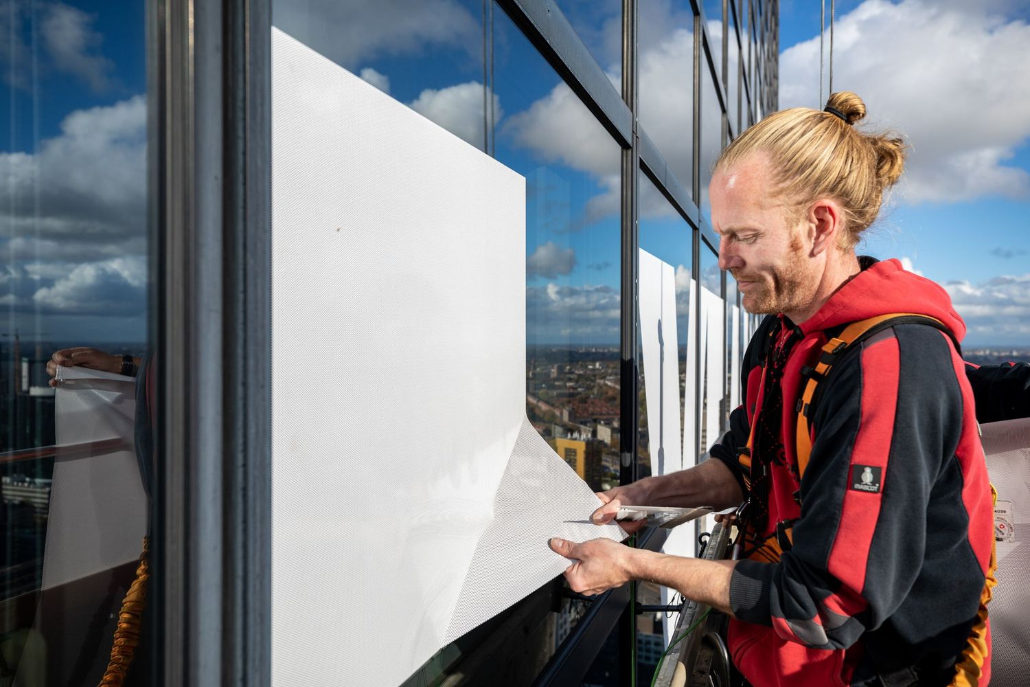 Man with bun hairstyle installing white panel on high-rise building window with city skyline visible in background.