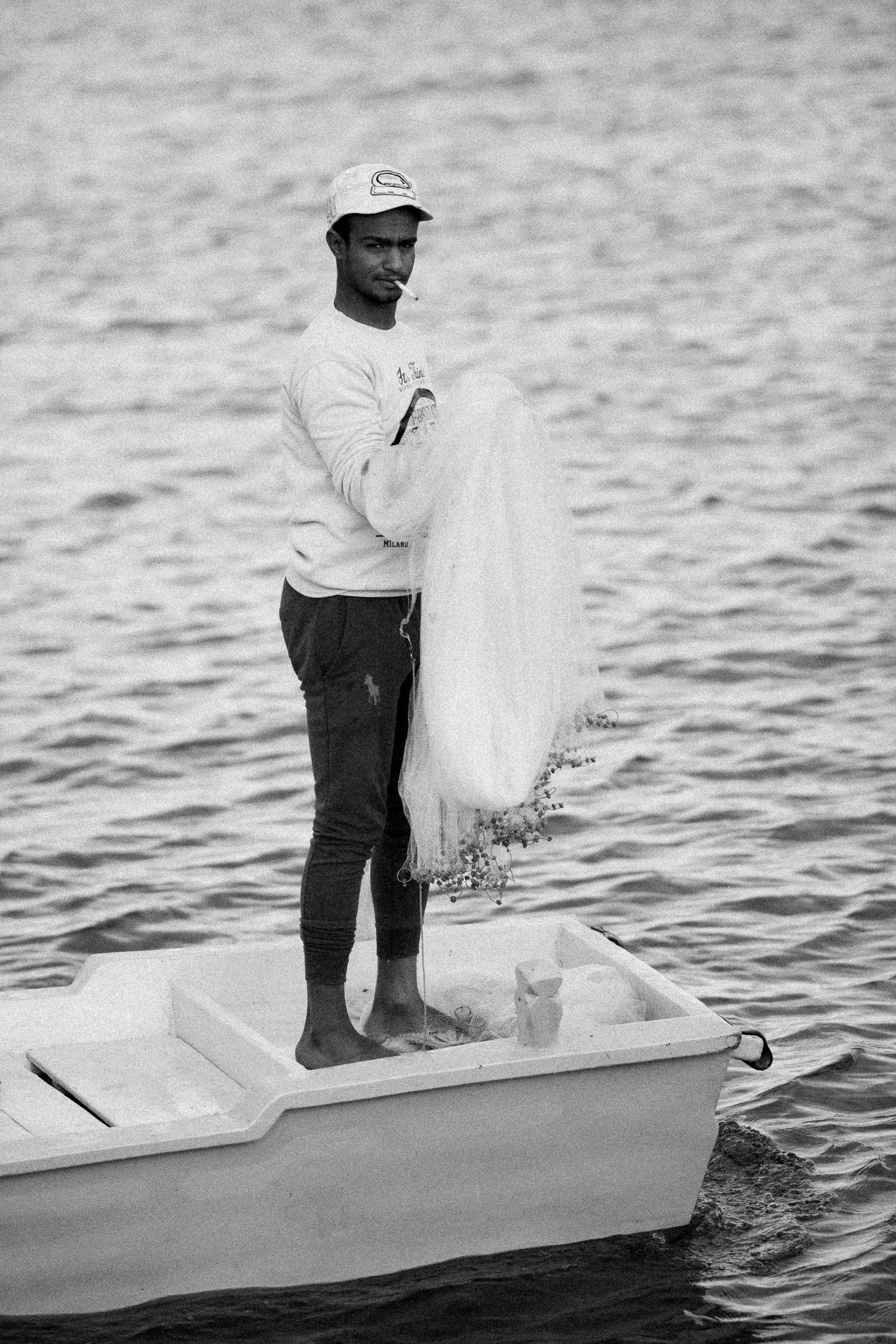 Black and white photo of a fisherman throwing a net from a small boat on calm waters.