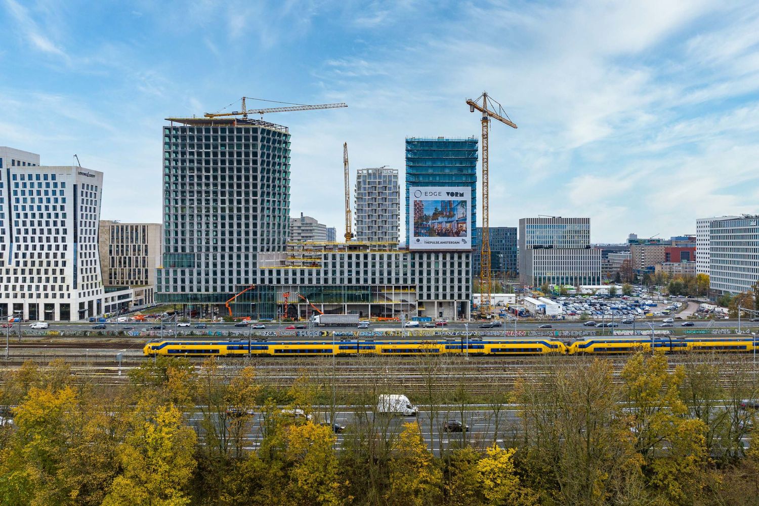 Urban skyline with construction cranes, modern buildings, yellow train, and autumn trees under a blue sky.