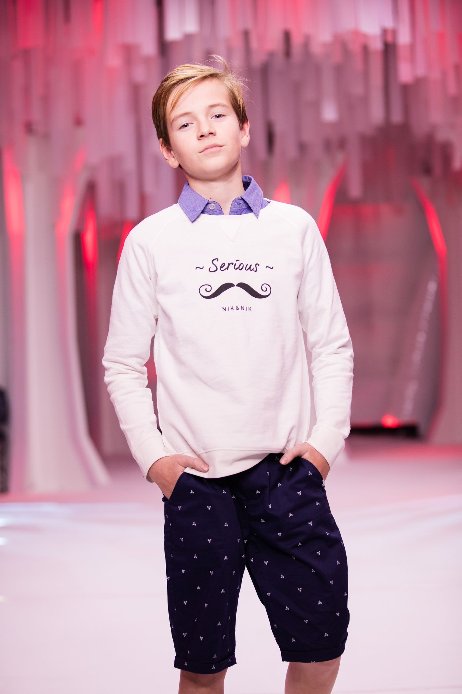 Boy model wearing a white Serious sweatshirt over a blue shirt with navy patterned shorts on a runway.