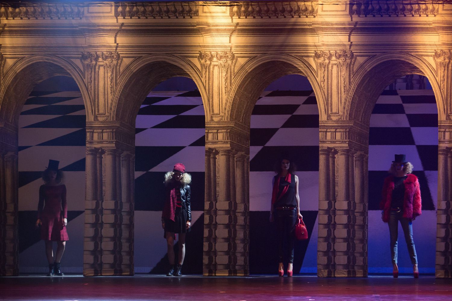 Performers in red costumes stand beneath ornate golden arches with geometric black and white backdrops on a theatrical stage.
