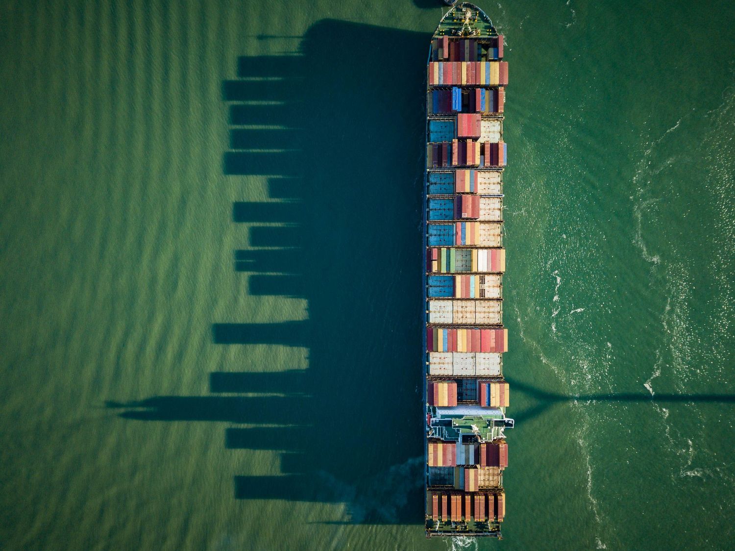 Aerial view of a cargo container ship with colorful containers casting long shadows on green ocean waters.