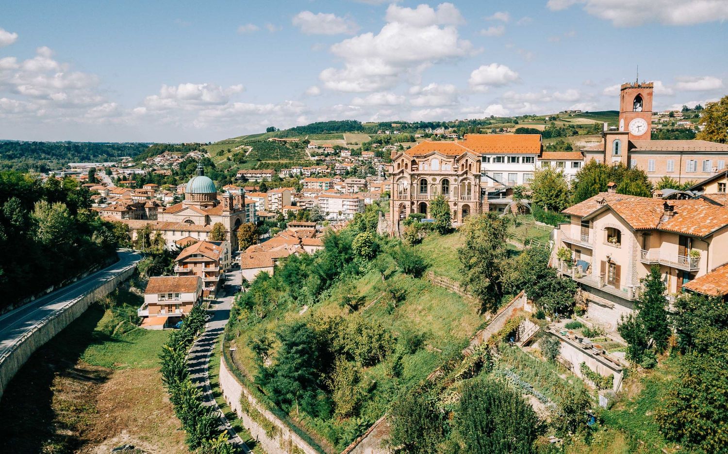 Aerial view of a charming Italian hillside town with terracotta rooftops, a church tower, and lush greenery under a partly cloudy sky.
