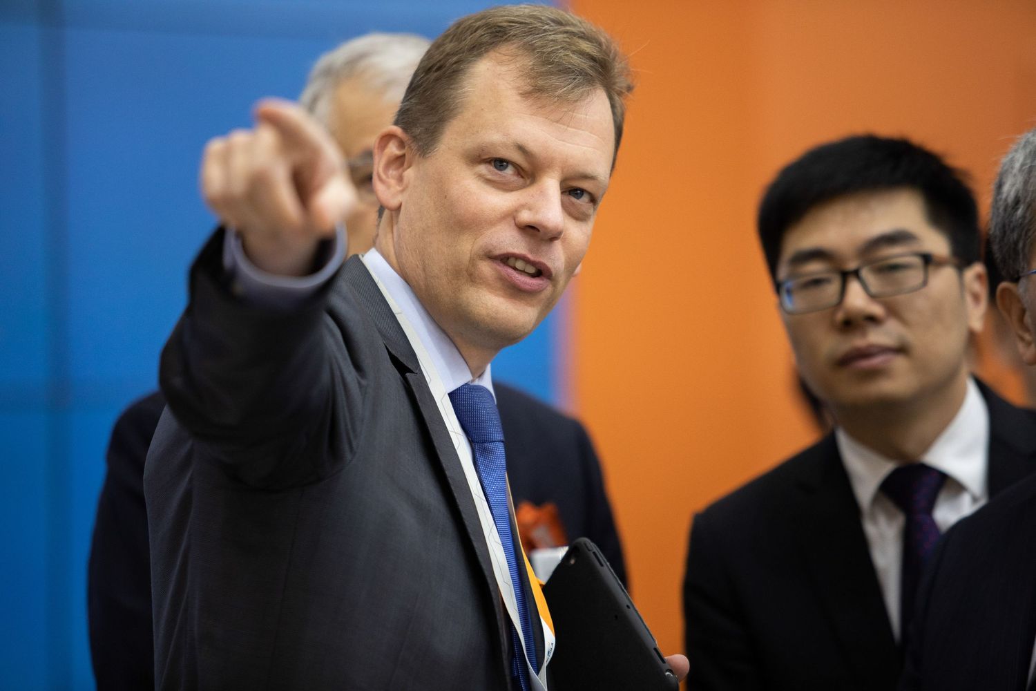 A person in a suit points forward while standing next to another person in front of orange and blue walls.