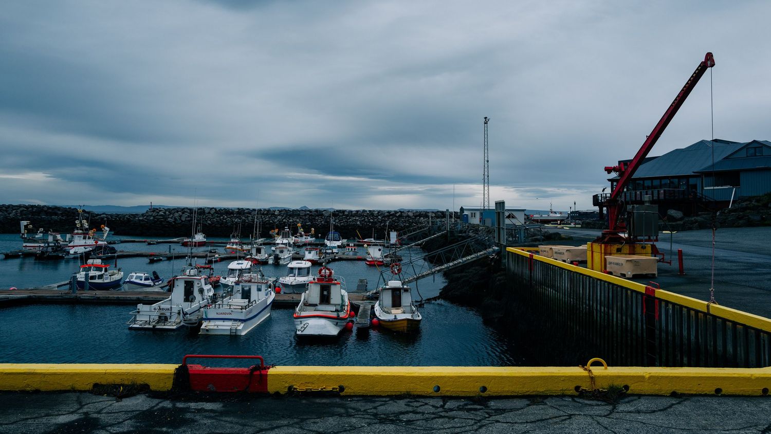 Boats docked at a marina with industrial cranes and cloudy skies in the background.