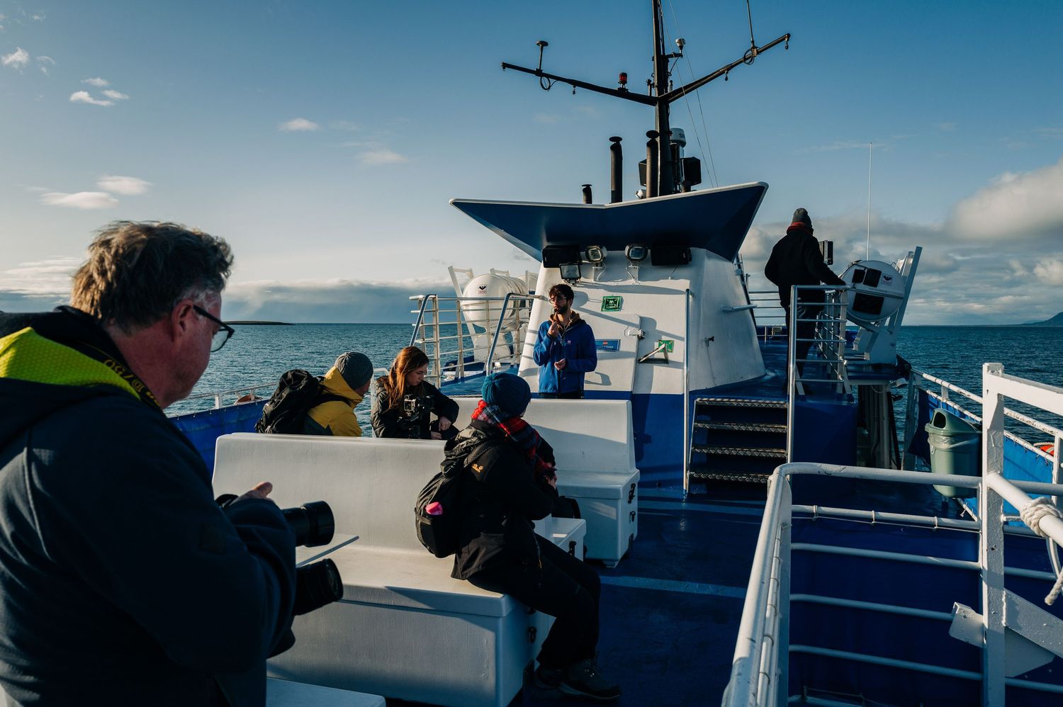 Passengers enjoy views from the deck of a blue and white ferry boat sailing across calm waters on a sunny day.