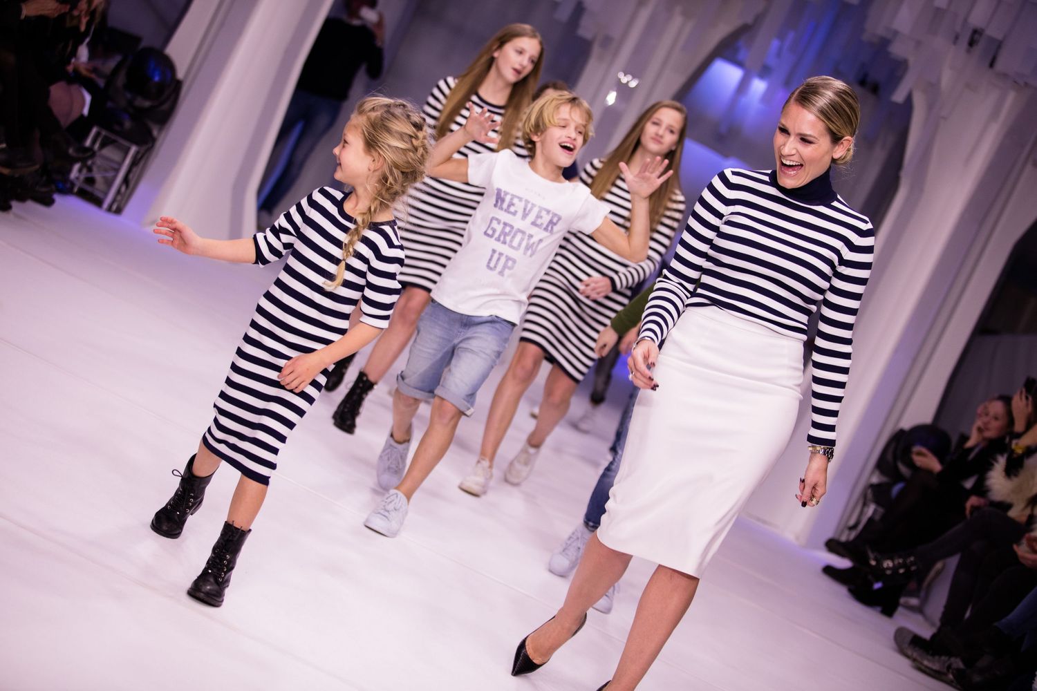 Women and girls in striped tops walk a fashion runway, smiling and dancing together at a children's fashion show.