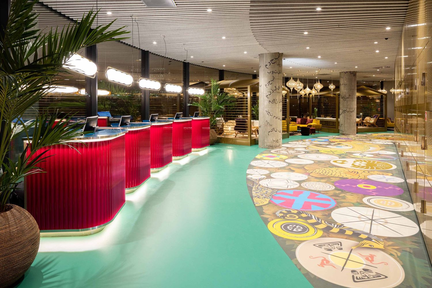 Modern restaurant interior with red booth seating and colorful circular floor patterns alongside decorative palm plants.