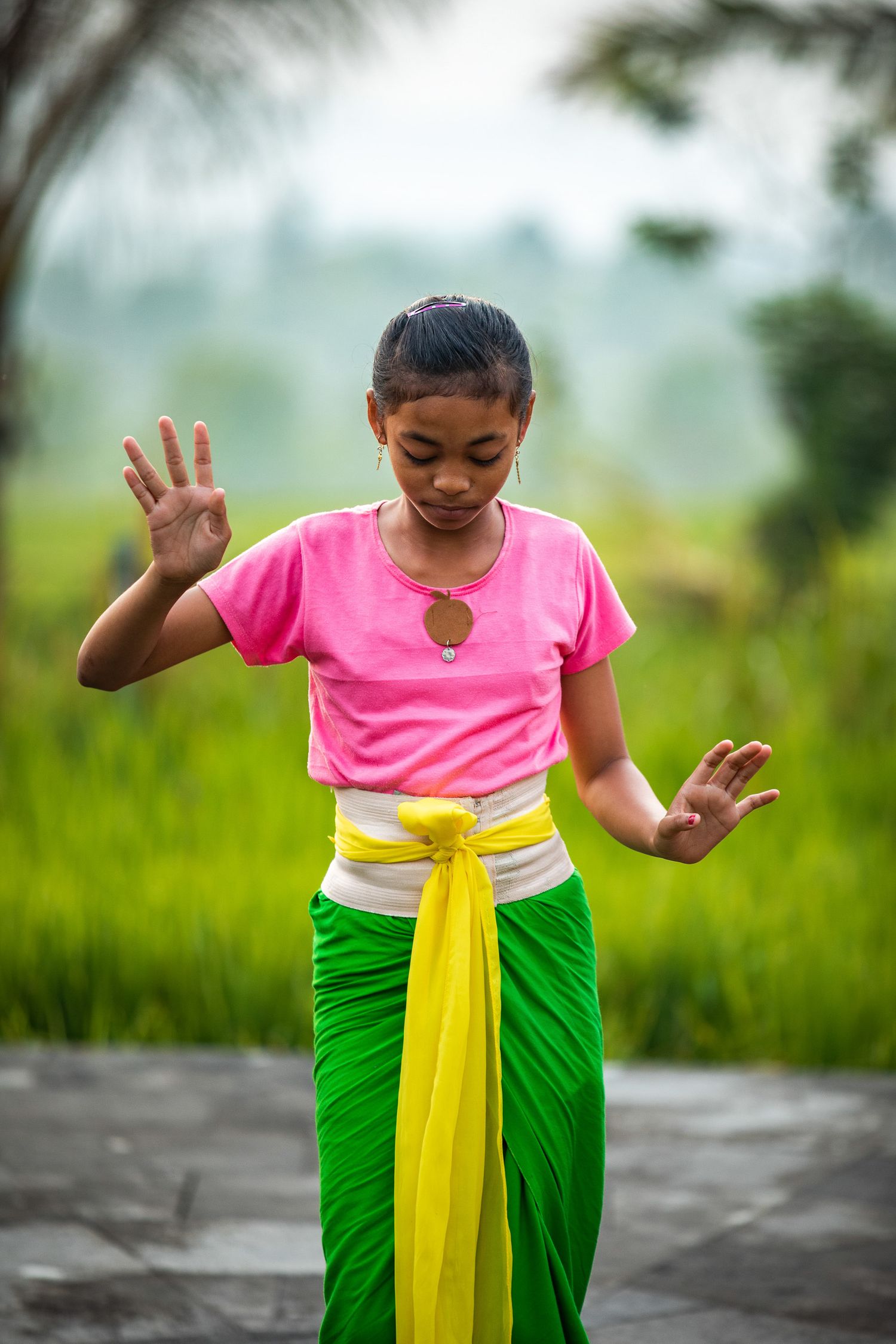 Young dancer in traditional Balinese costume with pink top, green skirt and yellow sash performs graceful movements.