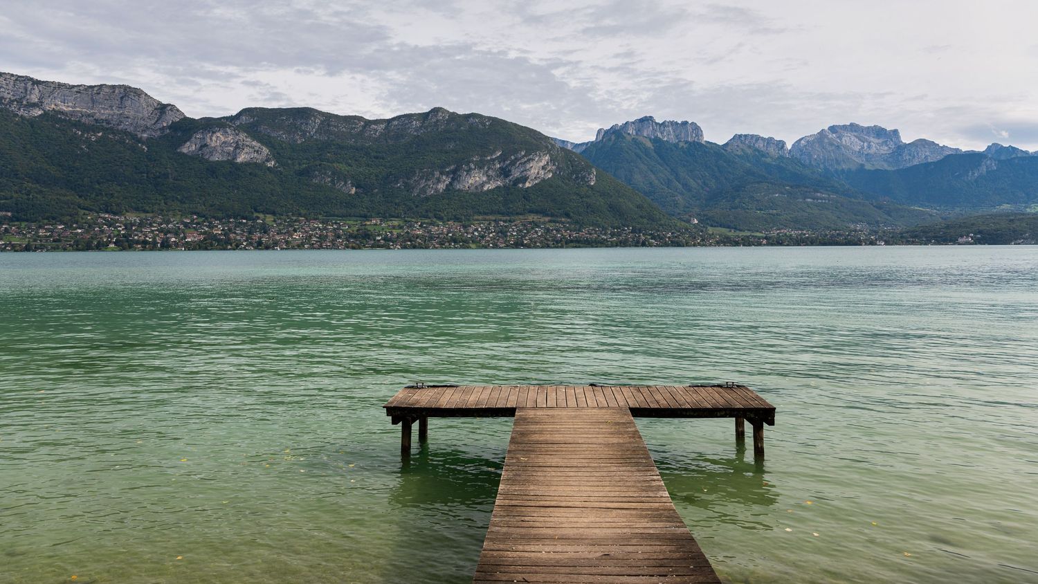 Wooden pier extends into tranquil green lake waters with mountains in the background.