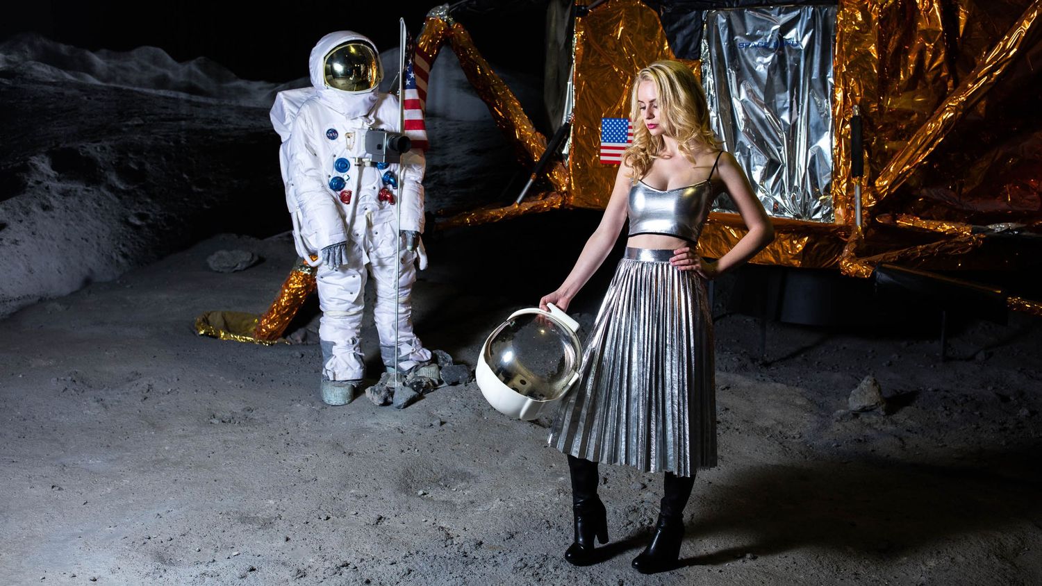 Woman in silver dress holds astronaut helmet beside spacesuit figure near lunar lander with American flag backdrop.