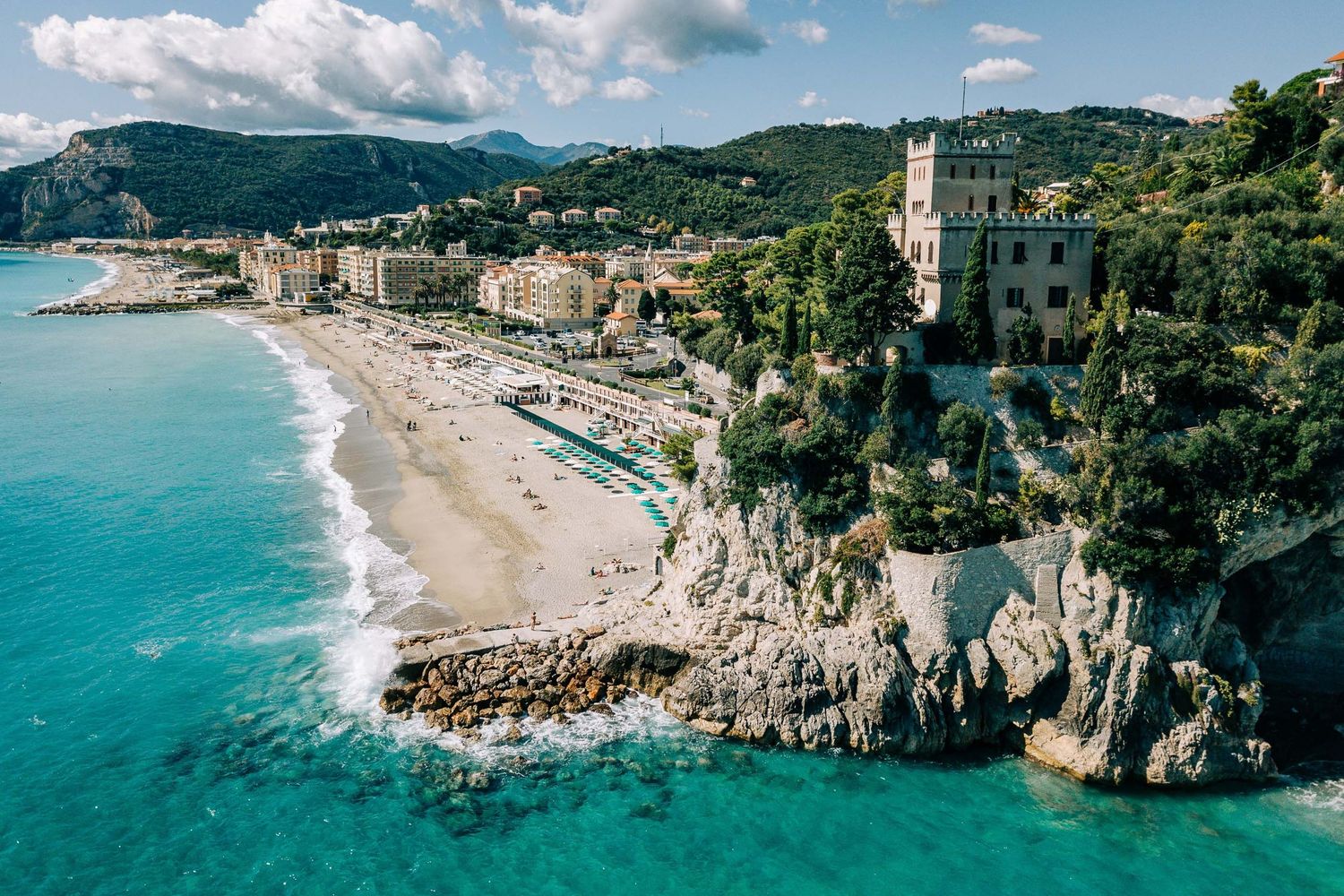 Aerial view of a scenic Italian coastal town with turquoise waters, sandy beach, and cliffside castle.
