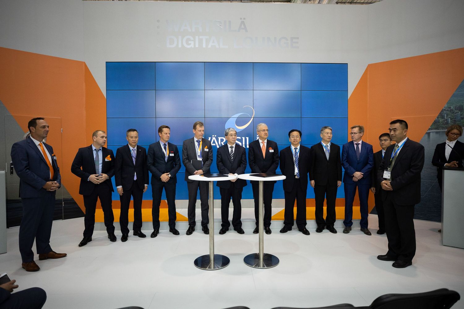Business professionals in dark suits stand in a modern office space with bright blue and orange walls and white tables.
