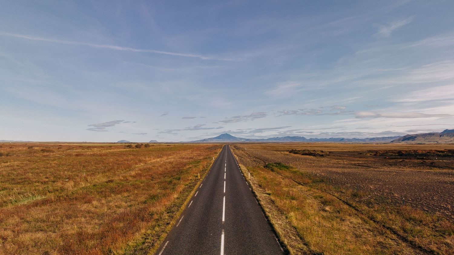 Long empty road stretches through vast brown plains with mountains in the distance.