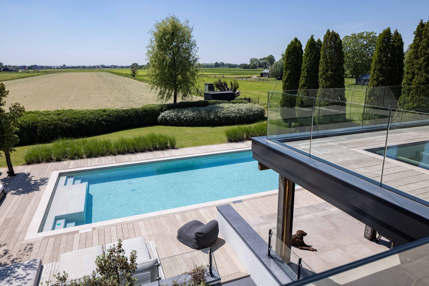 Modern backyard with a rectangular swimming pool, wooden deck and glass railings overlooking rural countryside views.