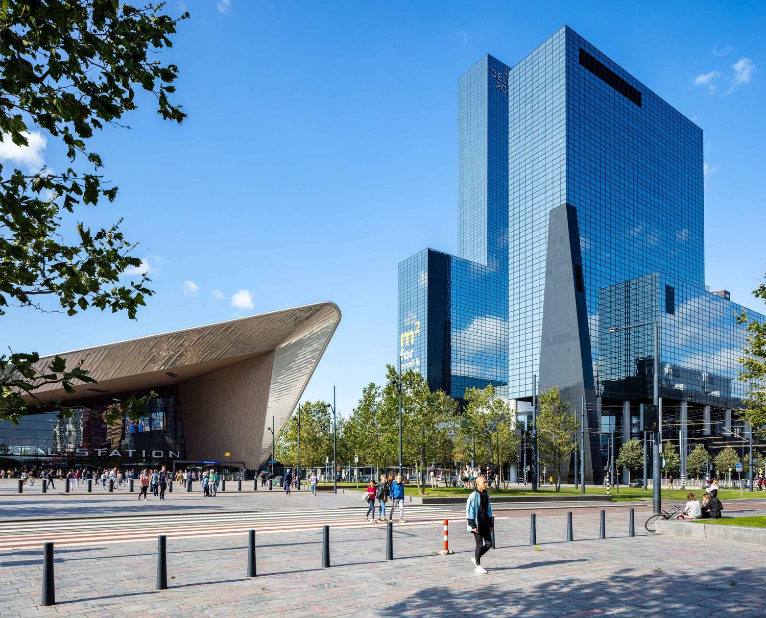 Modern glass office buildings and architectural structures line an urban plaza under bright blue skies in Rotterdam.