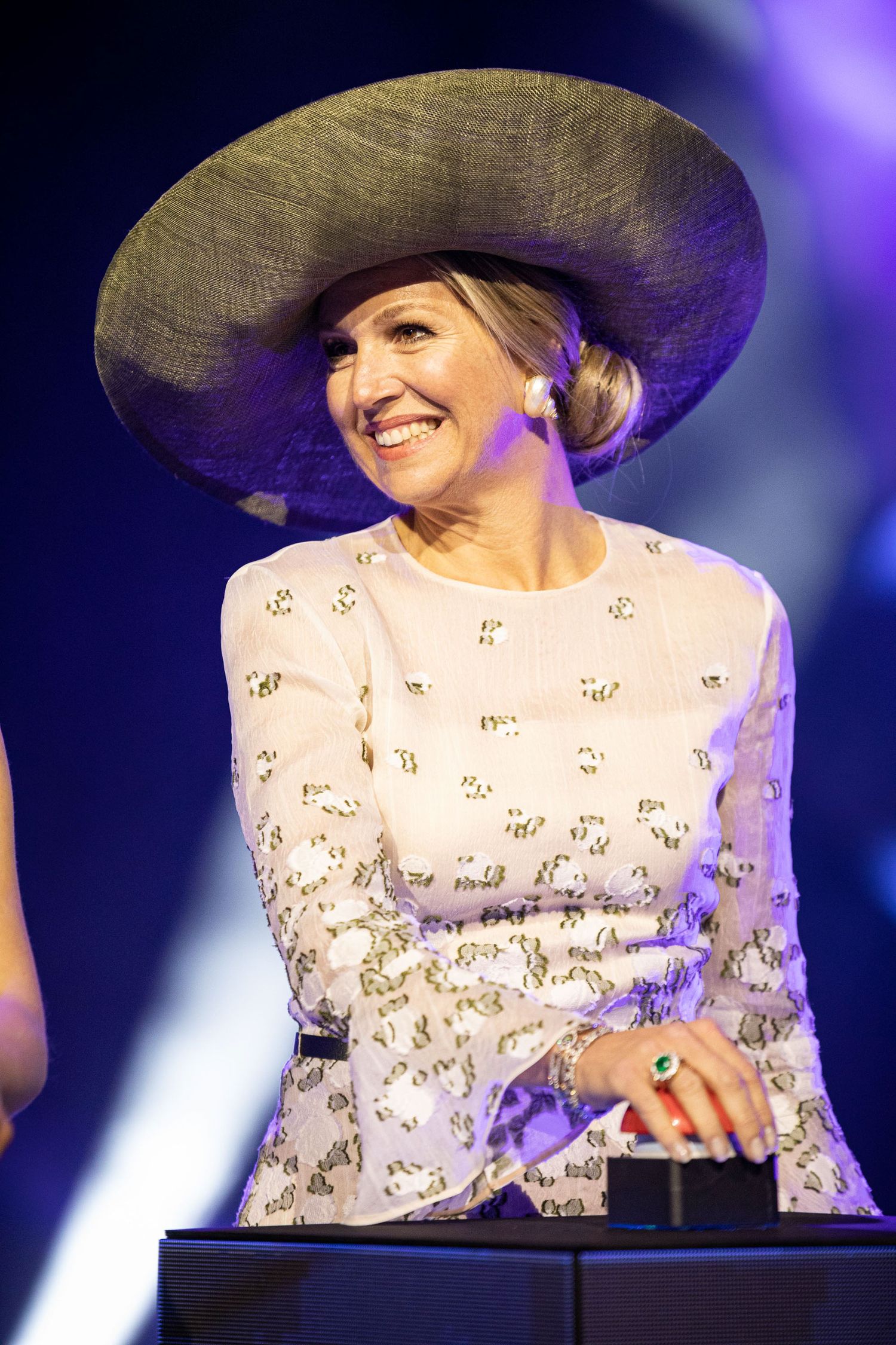 A performer in a wide-brimmed hat and white lace dress smiles while on stage under purple lighting.