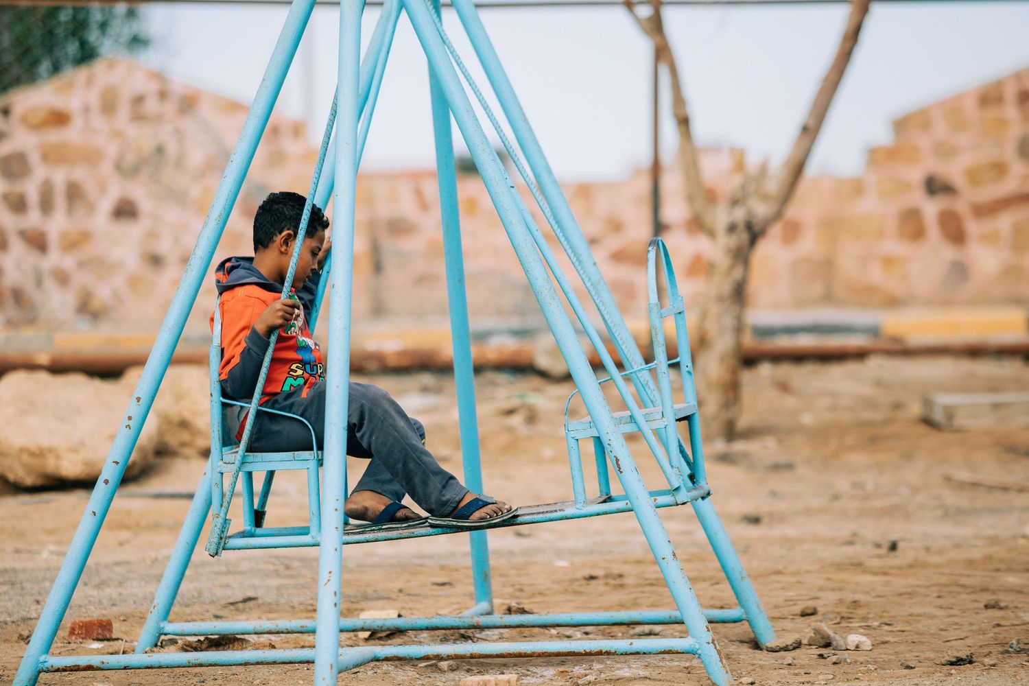 A blue metal swing set stands against a desert landscape with brick buildings in the background.