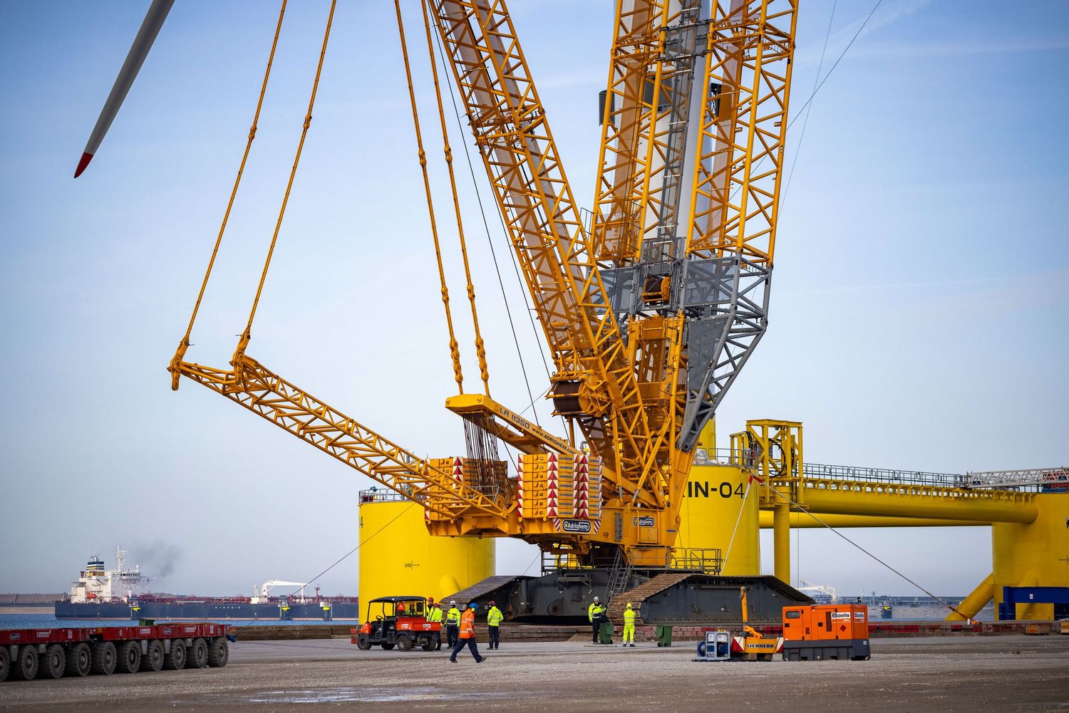 Heavy duty cranes lifting massive yellow wind turbine components at a port.