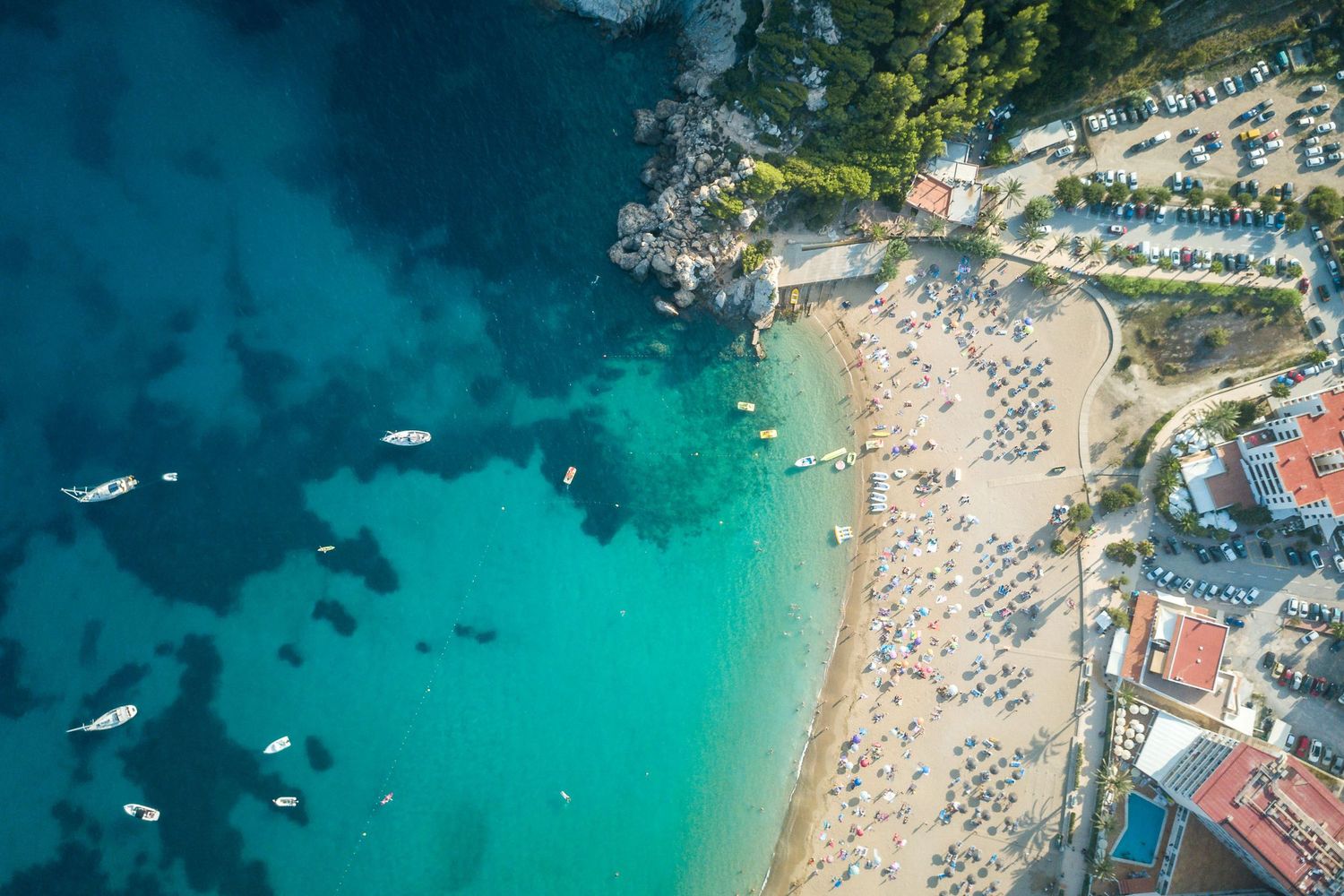 Aerial view of a stunning turquoise bay beach with boats, sunbathers, and lush green cliffs in a Mediterranean resort town.