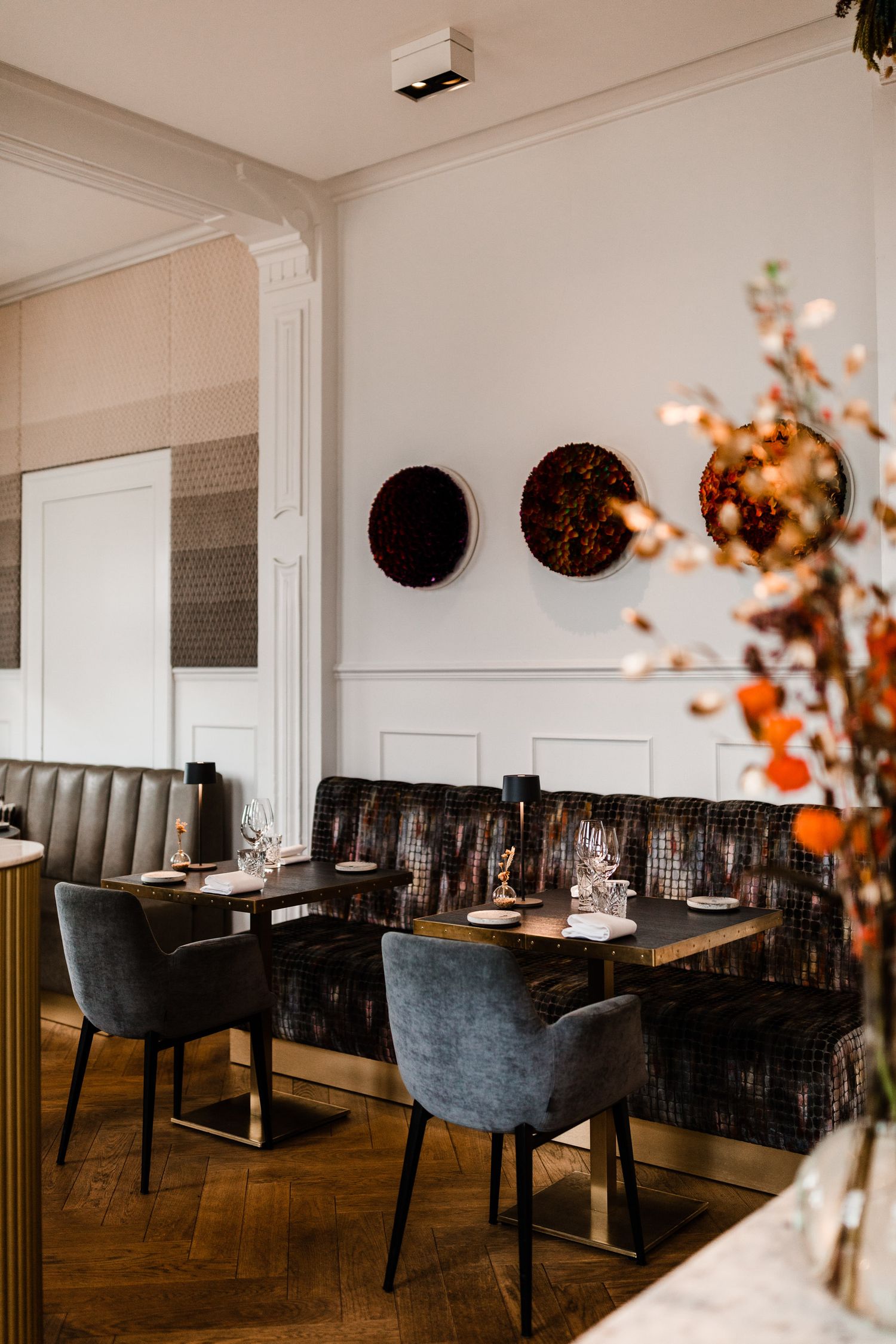 Modern restaurant interior with dark upholstered seating, white walls, and autumn floral arrangements in warm lighting.