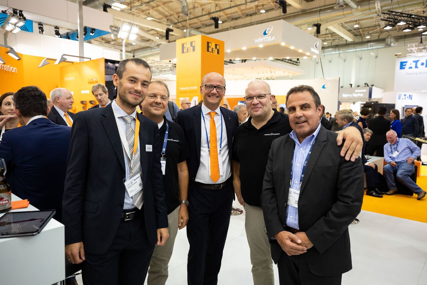 Business professionals in suits gather together for a photo at a brightly lit trade show or convention center.