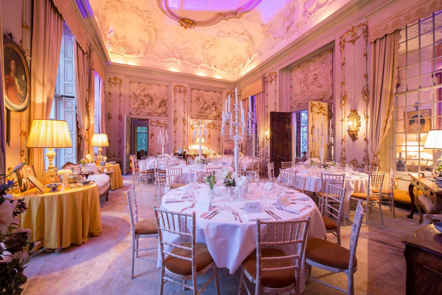 Elegant baroque banquet hall with ornate frescoed ceiling, round tables set for dining, and golden ambient lighting.