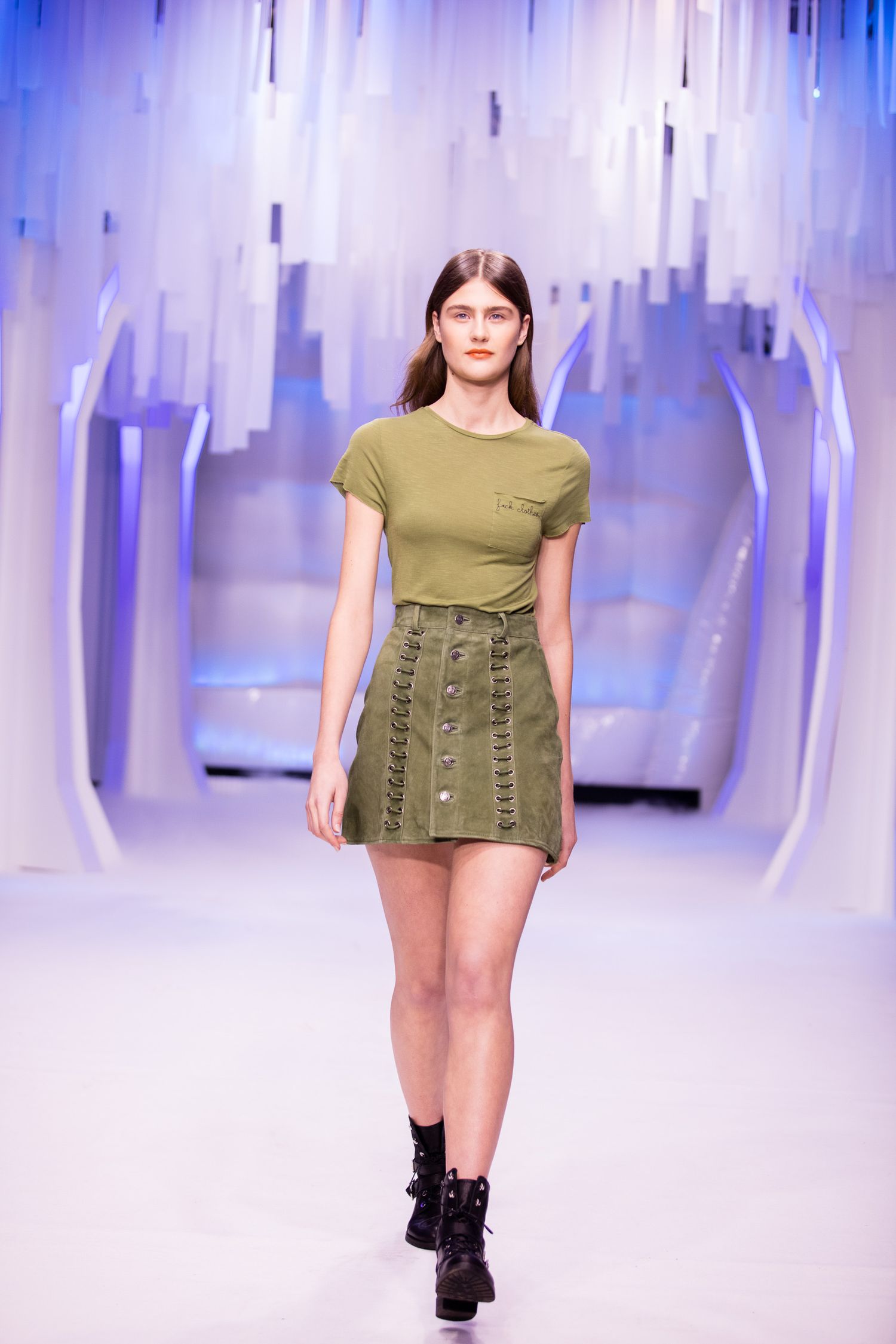 Model walks runway in olive green t-shirt and matching mini skirt with black boots at fashion show.