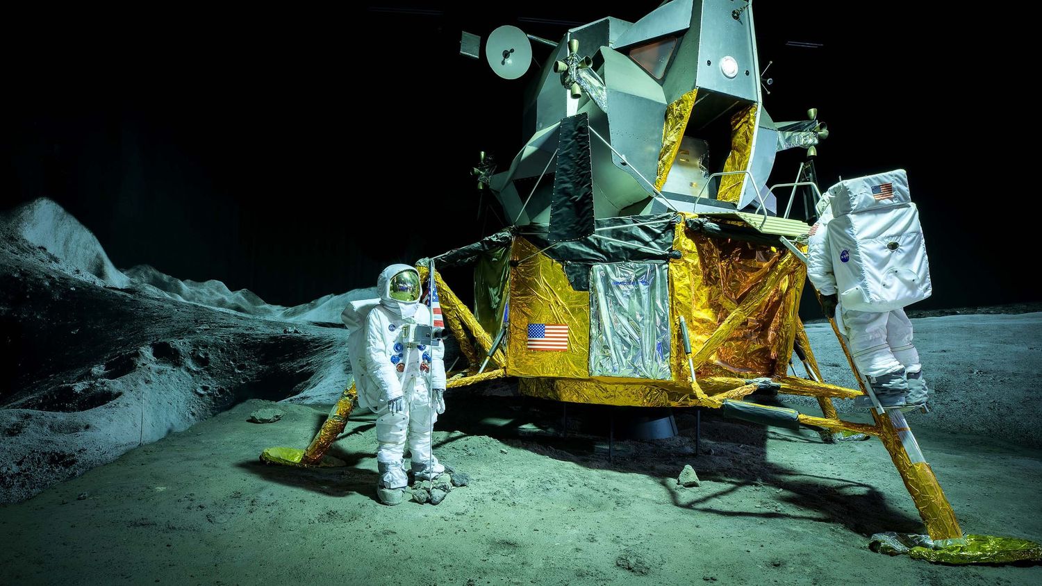 Two astronauts in white spacesuits stand beside a yellow lunar lander module on a simulated moon surface.