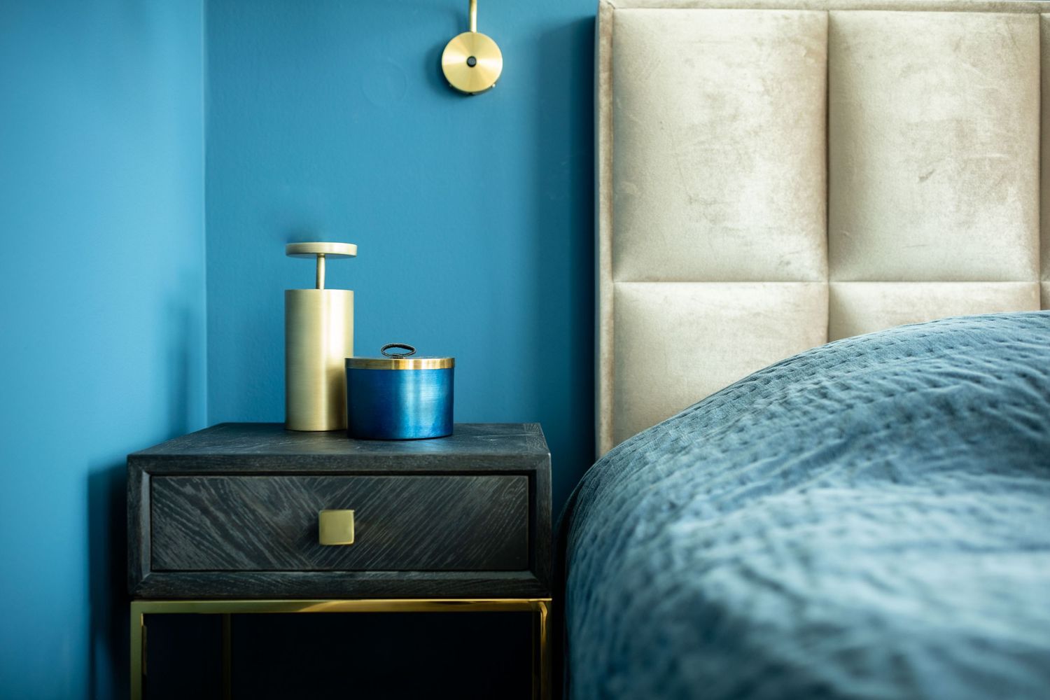 Modern bedroom interior with teal blue wall, beige headboard, and black nightstand with decorative containers.
