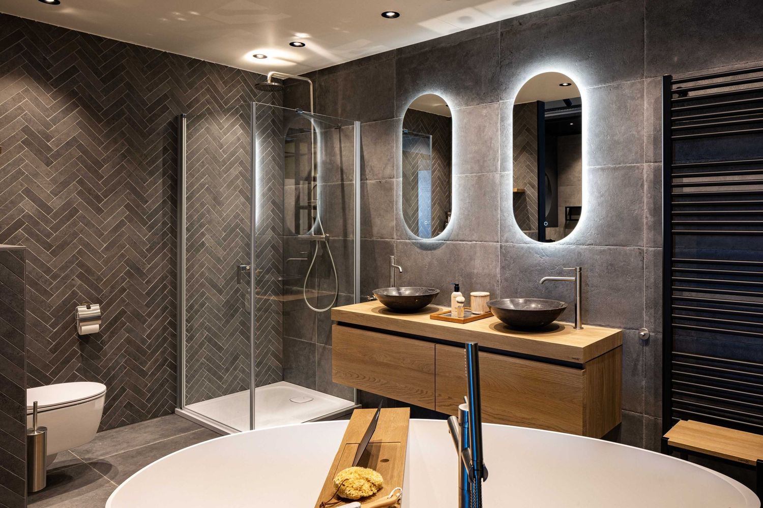 Modern bathroom interior with gray herringbone tile walls, black fixtures, and oval mirrors creating a luxurious spa-like atmosphere.