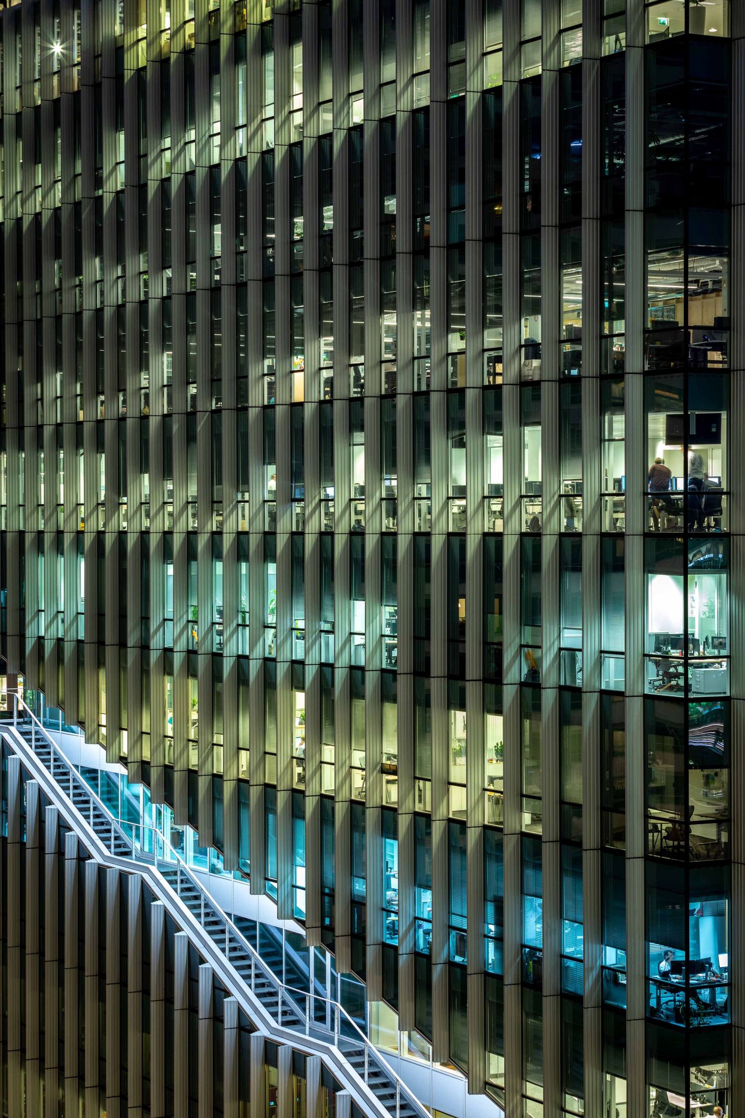 Modern office building at night with illuminated escalators glowing blue against dark glass exterior.