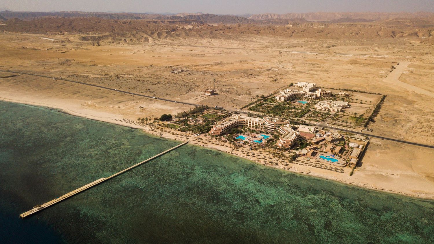 Aerial view of a luxury desert resort with pools and lush gardens along a coral reef coastline.