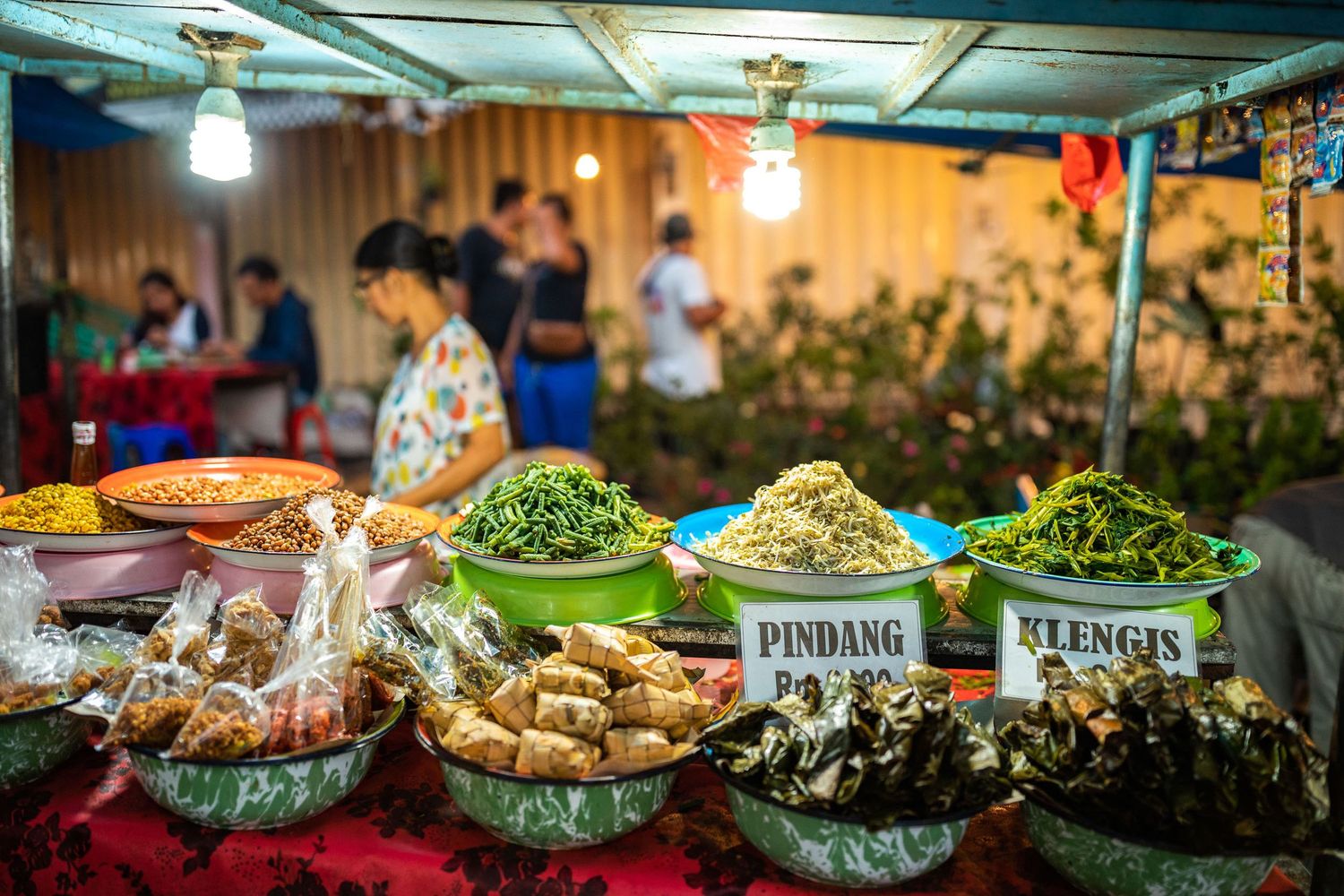 Various local dishes and food items displayed at an evening market under warm lighting.