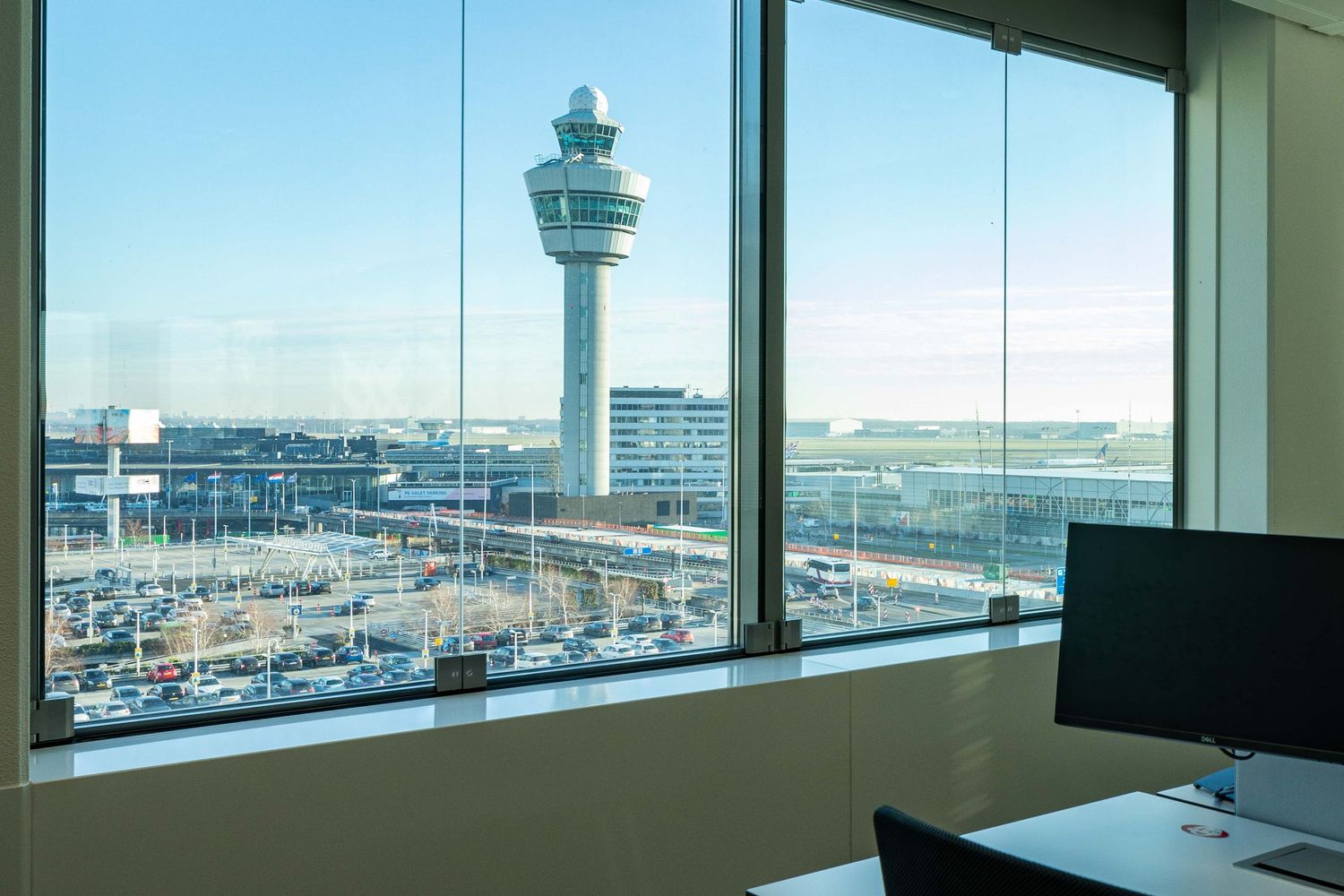 View through office window showing airport control tower and terminal buildings at Amsterdam Schiphol Airport.