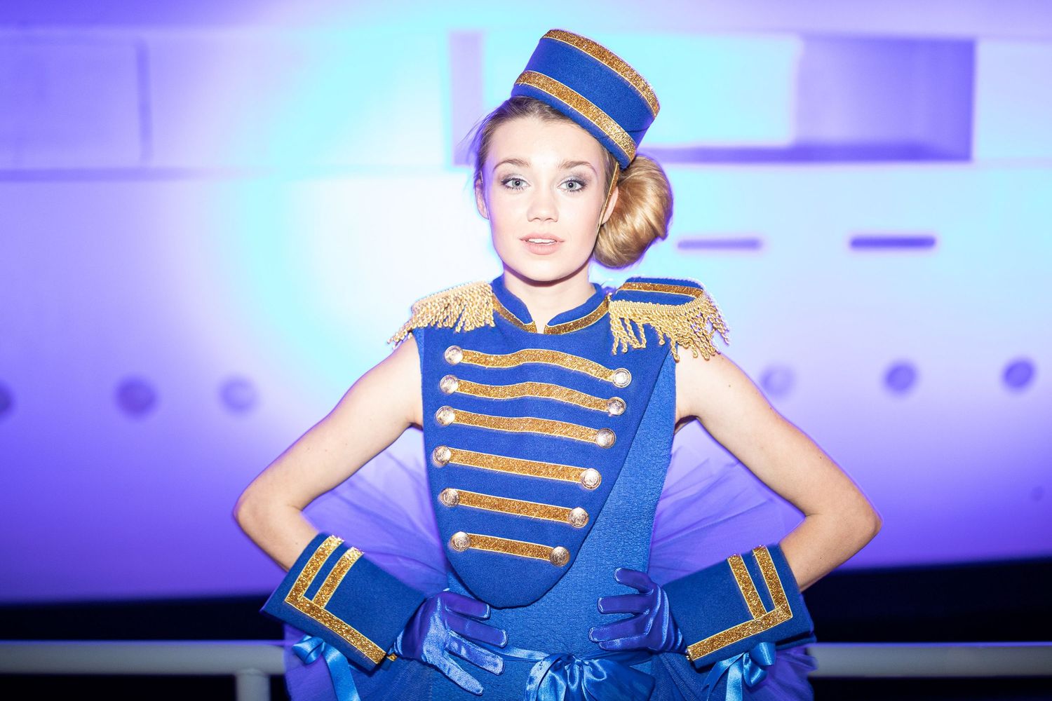 A performer in a blue and gold military-style costume dances on stage under purple lighting.