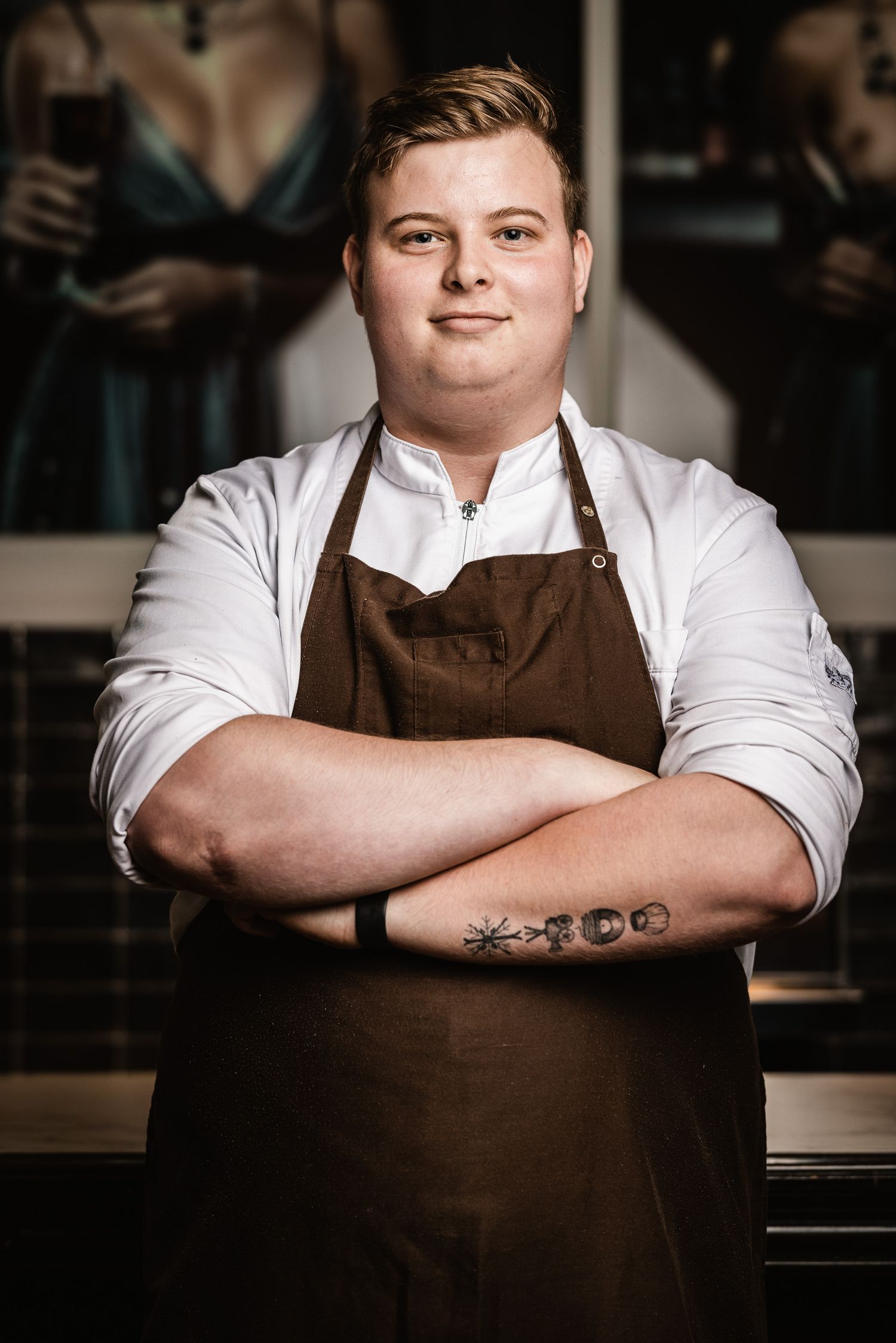 Professional chef wearing white shirt and brown apron stands confidently in restaurant kitchen with crossed arms.