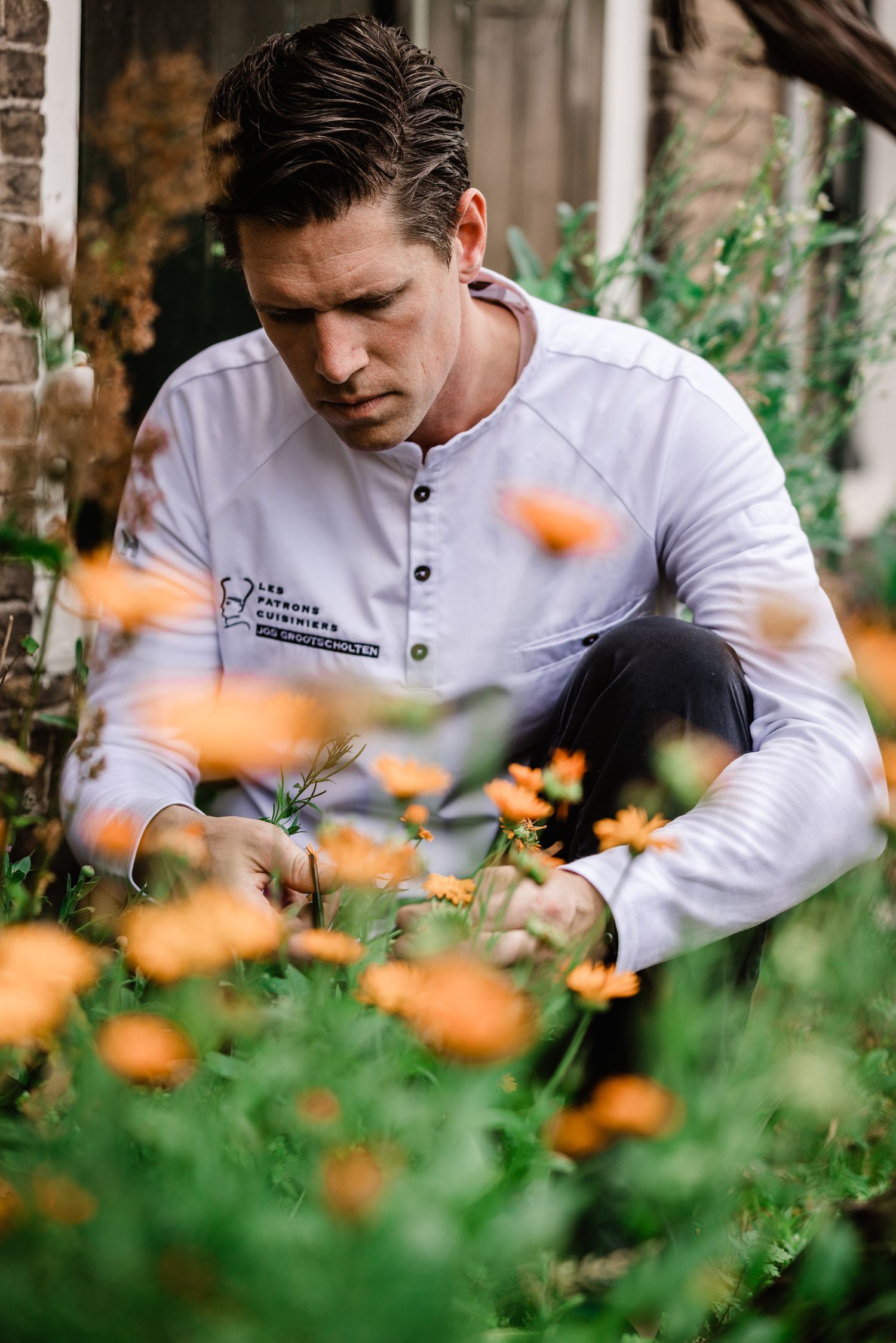 Chef in white uniform carefully examines herbs and flowers in a lush garden setting with orange blooms in foreground.