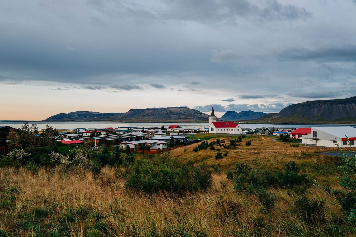 A panoramic view of red-roofed buildings nestled against dramatic mountains under cloudy skies in Iceland.