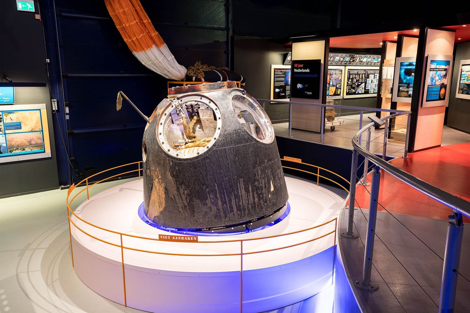 SpaceX Dragon spacecraft capsule on display in a museum exhibit with parachute visible overhead.
