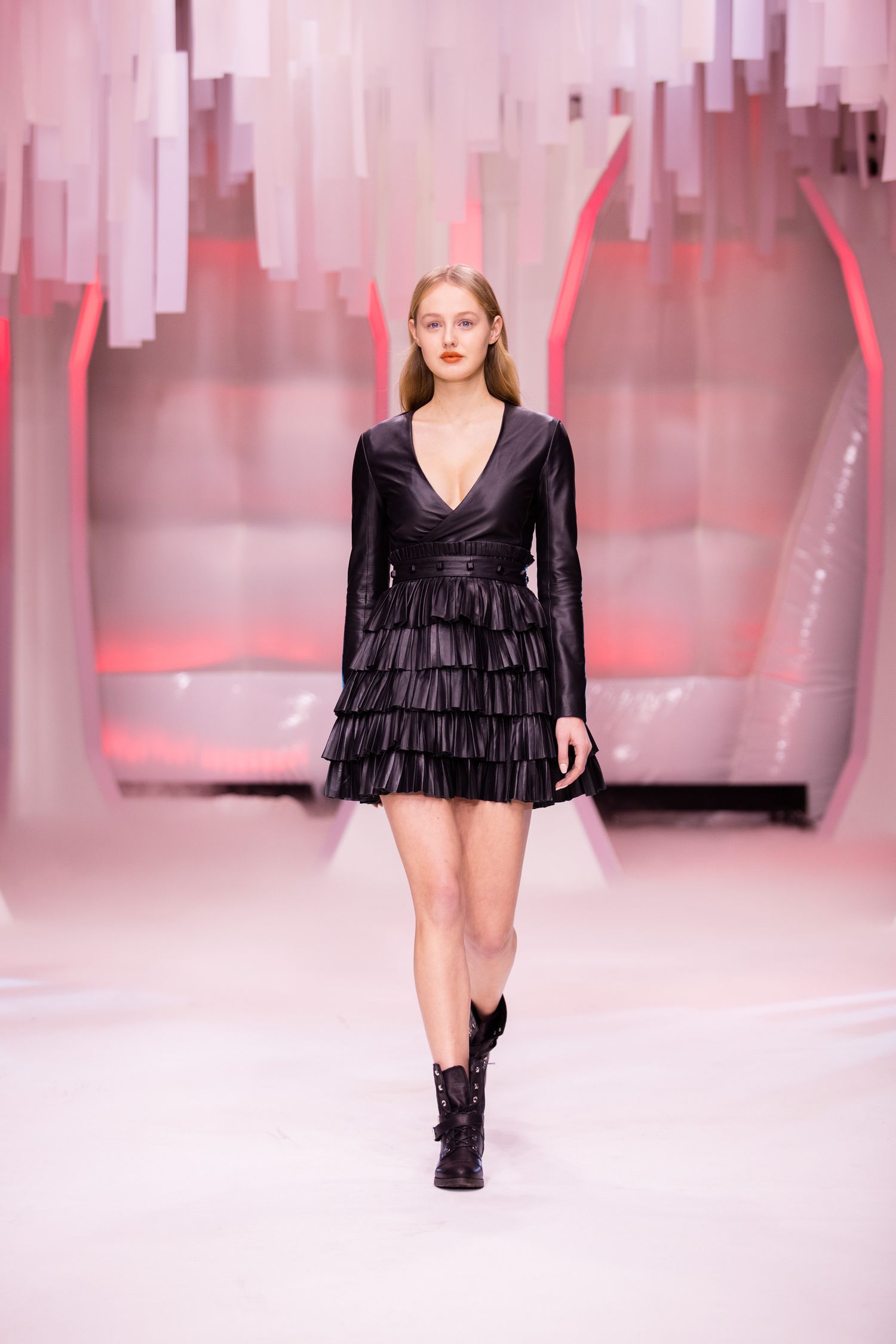 Model walks runway in black tiered ruffle mini dress with deep V-neck and ankle boots at fashion show.