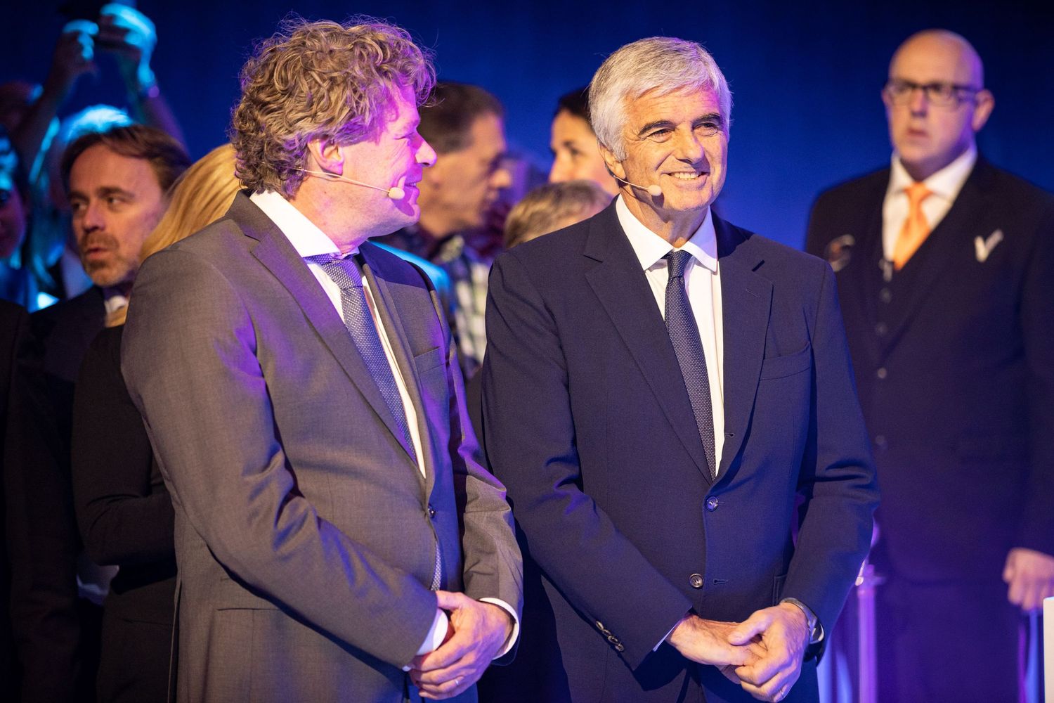 Two men in suits stand on stage at a formal event with blue lighting and additional people visible in background.